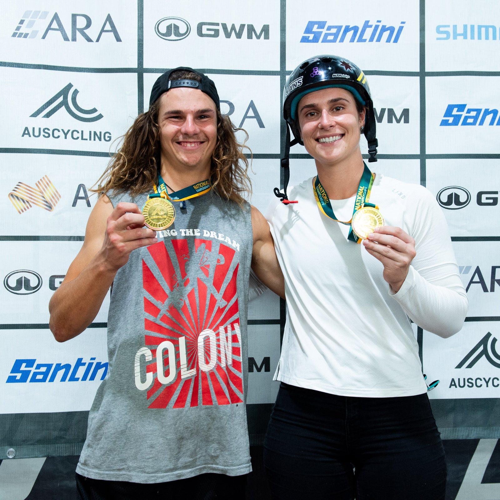 Two athletes holding gold medals in front of a branded backdrop